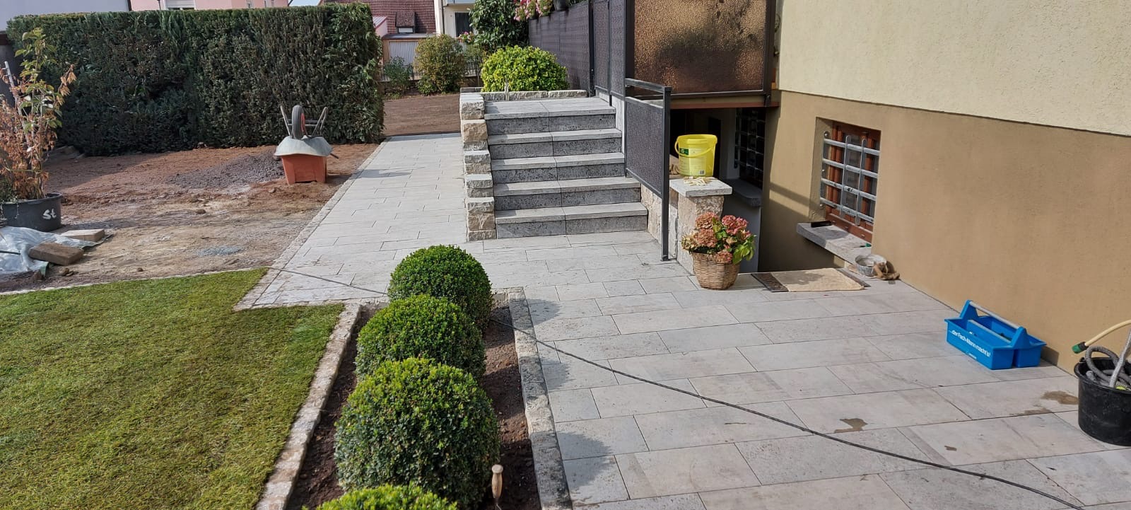 Jasnoszara kostka brukowa tworzy ścieżkę i schody w ogrodzie. Częściowo ułożony trawnik i rabaty z kulistymi krzewami. Widoczne narzędzia i materiały budowlane.