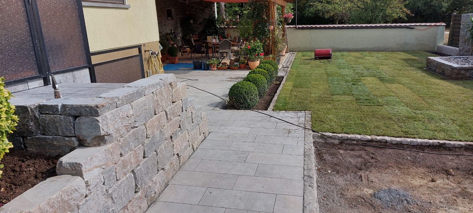 Nowy trawnik z rolki, obrzeża z kostki granitowej i ścieżka z płyt kamiennych w ogrodzie. Widoczna mur z kamienia łupanego.