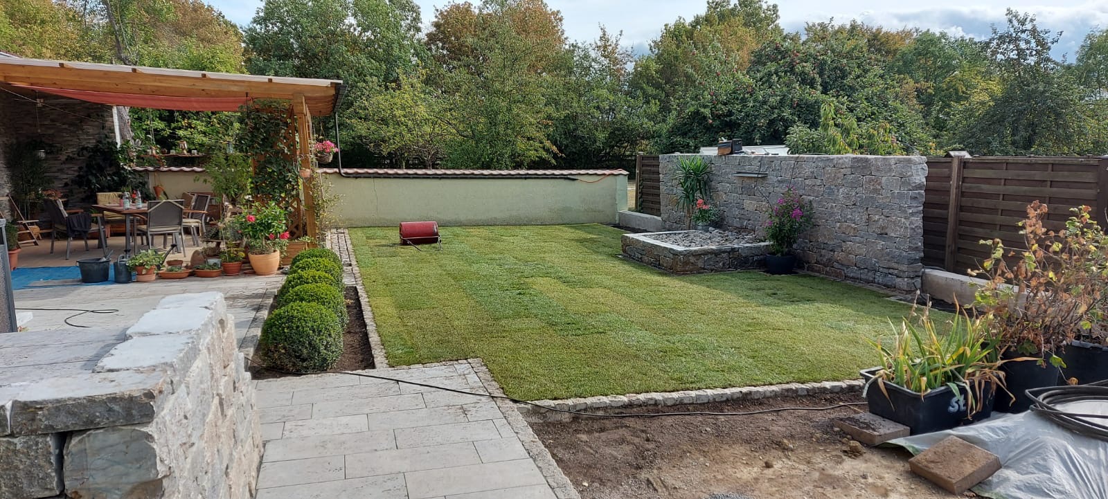 Zadbany trawnik z obrzeżem z kostki, fontanną z kamienia i altaną w tle. Widoczne rośliny w donicach i mała walcarka do trawy. Całość tworzy estetyczny ogród.