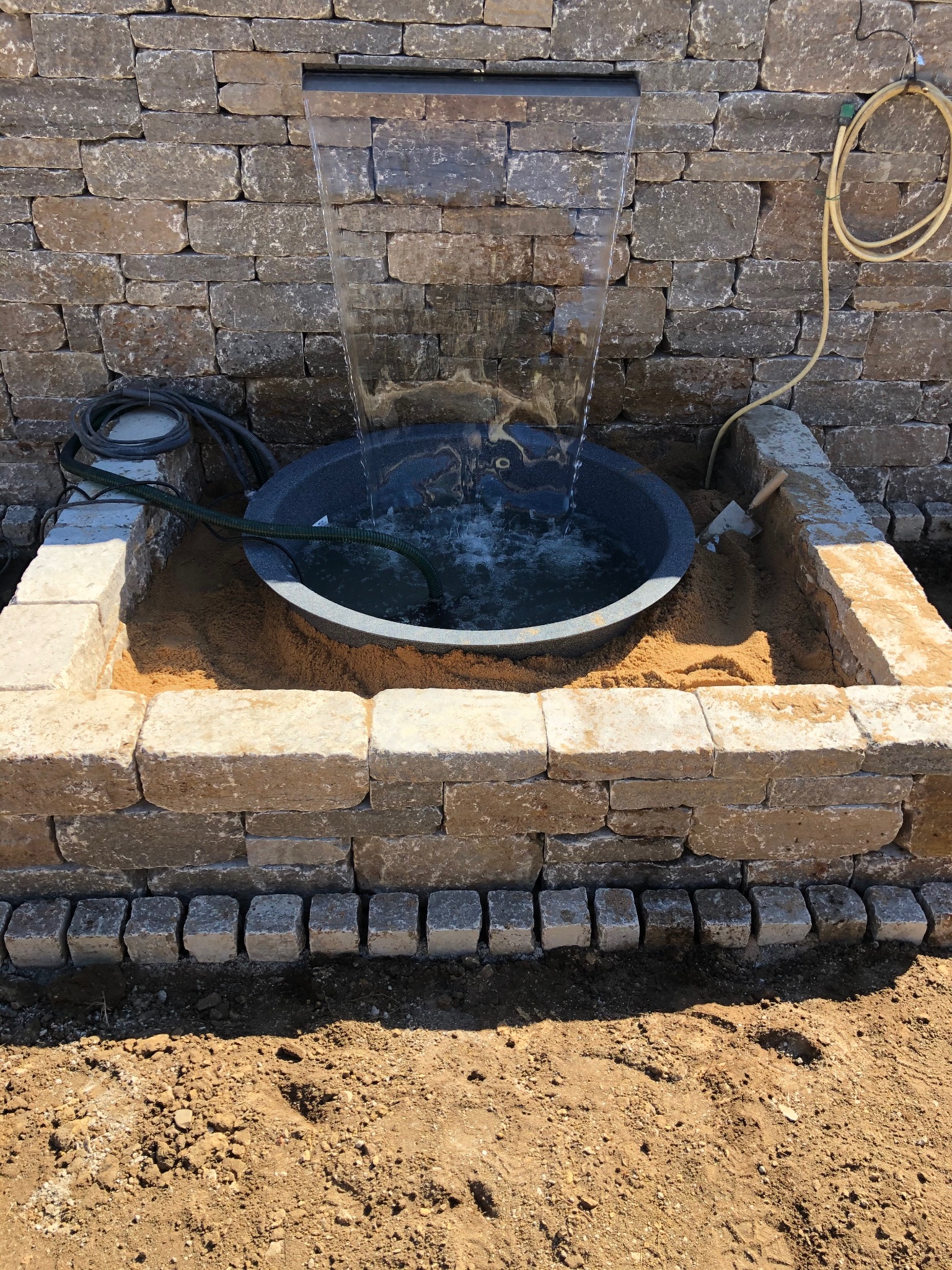 Fontanna z kamienia naturalnego w ogrodzie, otoczona kostką granitową. Elementy instalacji wodnej widoczne. Całość w surowym, naturalnym stylu.