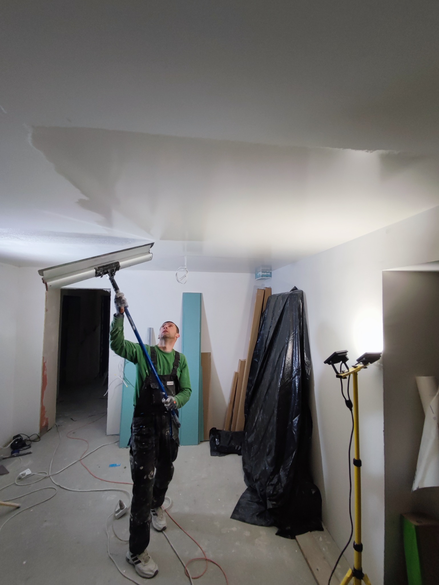 Pracownik w kombinezonie wygładza sufit za pomocą specjalistycznego narzędzia na wysięgniku. Widoczne niedoskonałości na suficie i elementy przygotowane do prac.