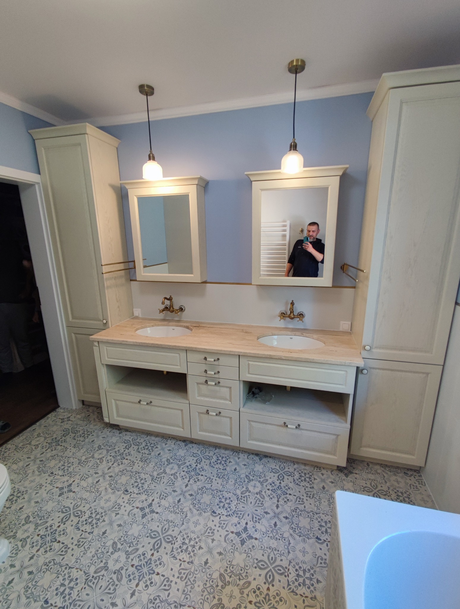 Elegancka, jasna łazienka w Warszawie z szafką pod umywalki, lustrami i wzorzystą podłogą. Widoczne baterie w stylu retro i oświetlenie.