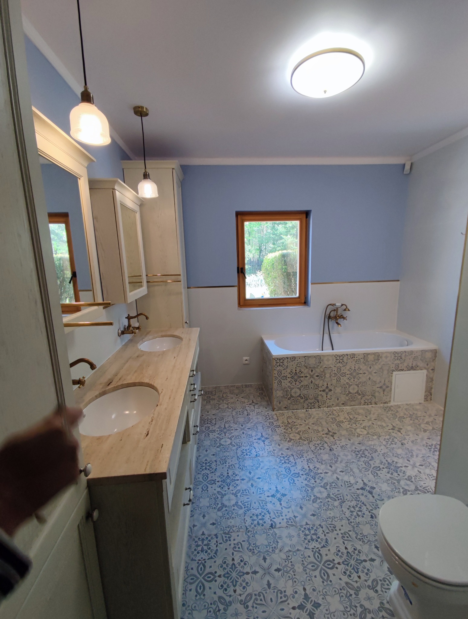 Jasna łazienka w stylu retro z drewnianymi akcentami: wzorzysta podłoga, wanna z obudową w mozaikę, umywalki nablatowe na blacie z jasnego kamienia.