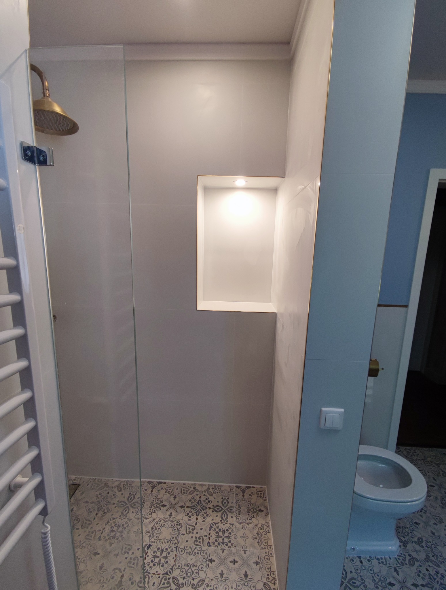 Wykończona łazienka: kabina prysznicowa z złotą deszczownicą, wnęka z oświetleniem, toaleta i wzorzyste płytki podłogowe. Detale z elementami złota.