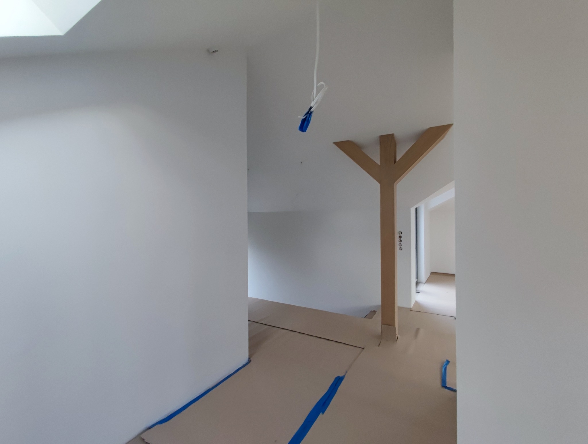 Białe ściany w trakcie prac wykończeniowych, zabezpieczona podłoga kartonem i taśmą malarską. Widoczny element konstrukcyjny w kształcie litery Y.