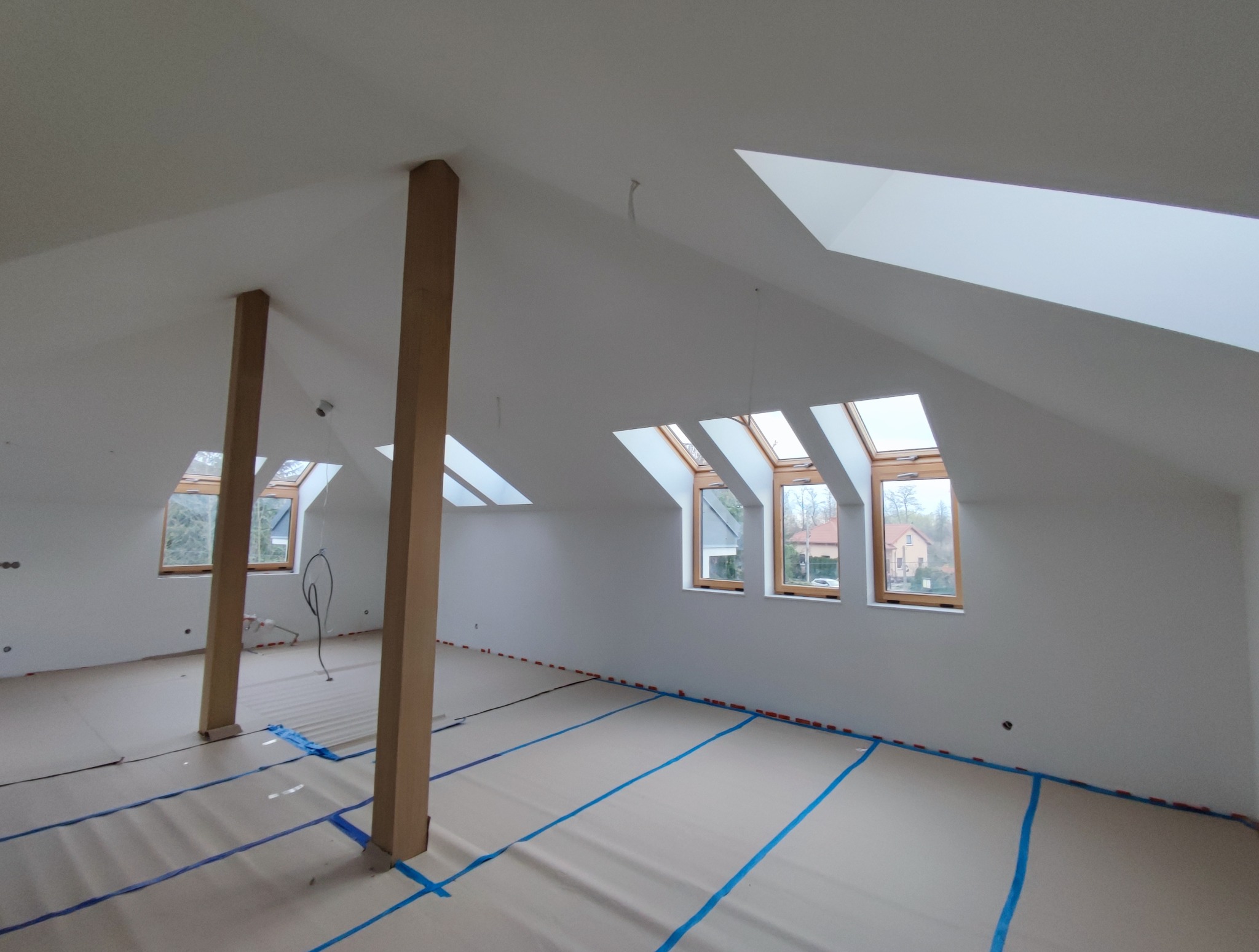 Poddasze po gładziowaniu i malowaniu na biało, z widocznymi oknami dachowymi i zabezpieczoną podłogą. Dwie drewniane kolumny podtrzymują strop.
