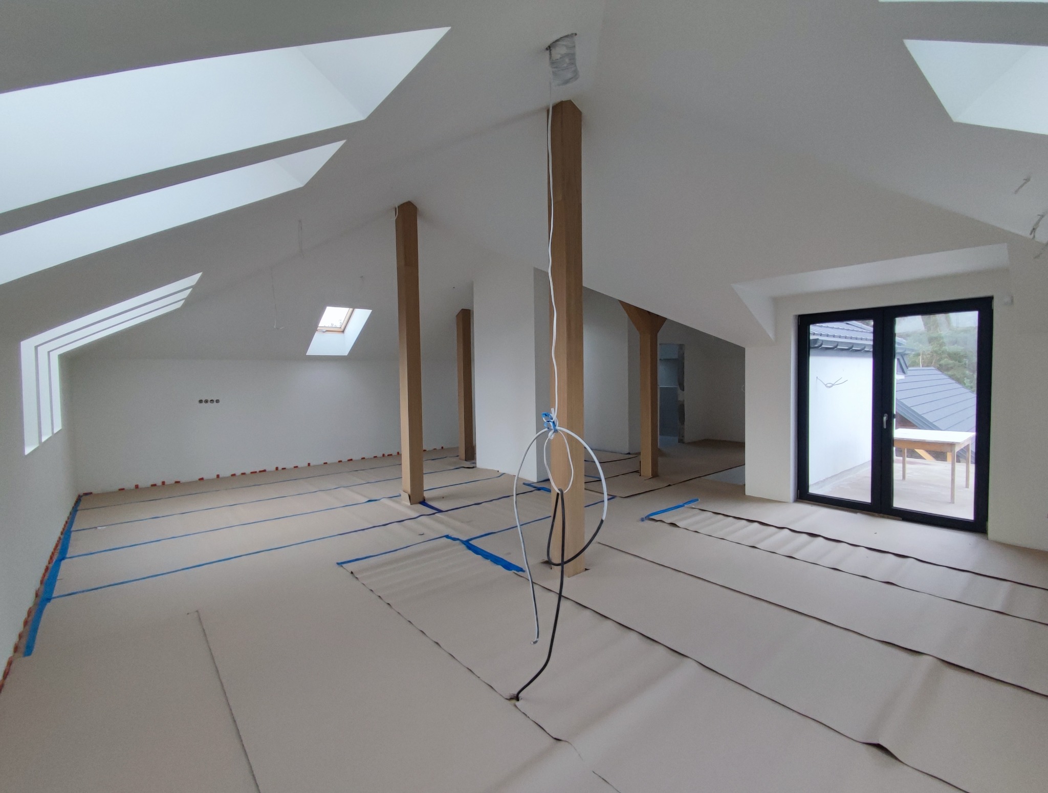 Poddasze w trakcie prac wykończeniowych: zabezpieczona podłoga, świeżo pomalowane ściany, drewniane słupy, okna dachowe i balkonowe. Widoczne kable elektryczne.