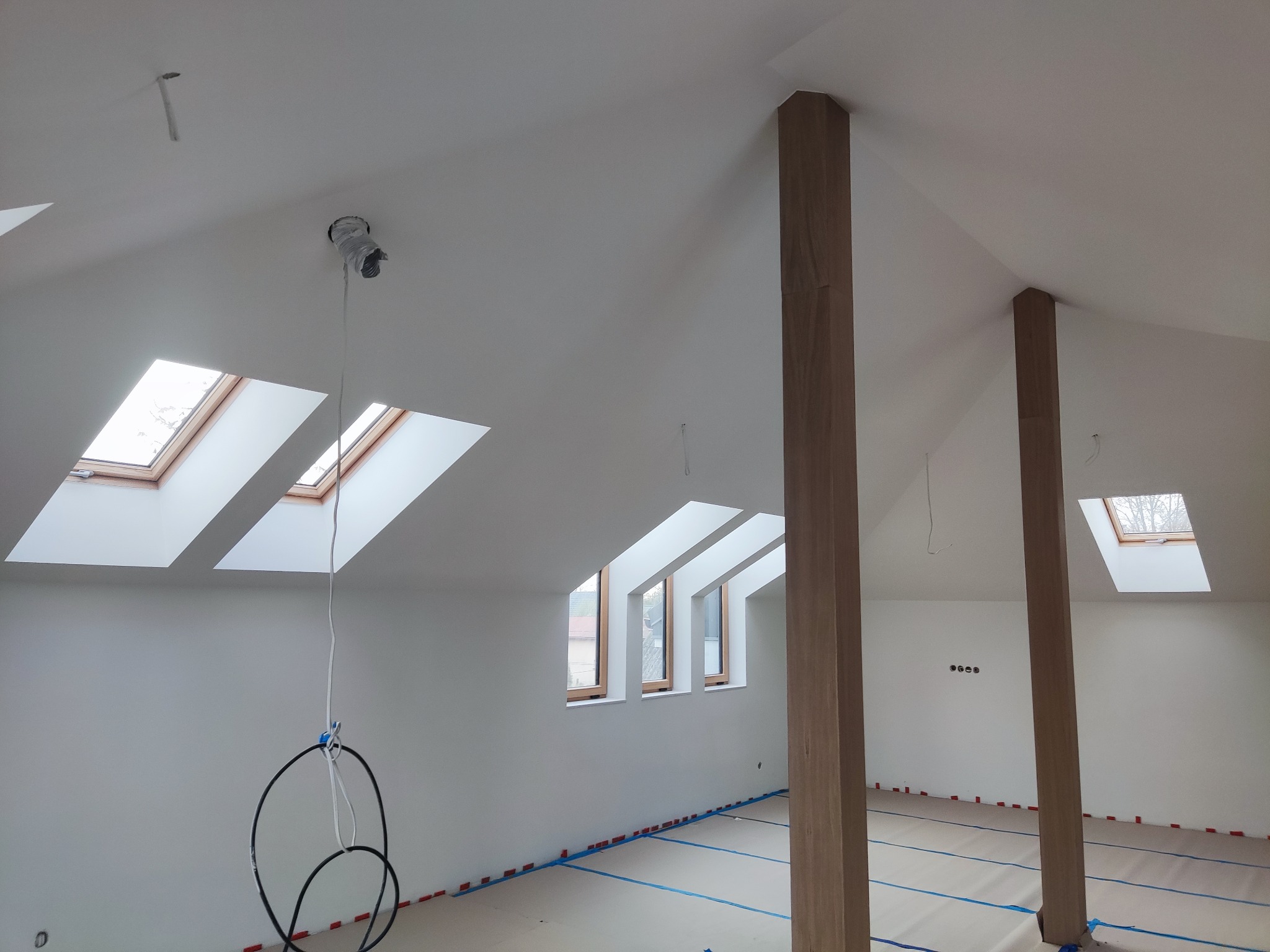 Poddasze po gładziowaniu i malowaniu na biało, z drewnianymi słupami i oknami dachowymi. Podłoga zabezpieczona folią, widoczne przewody oświetleniowe.