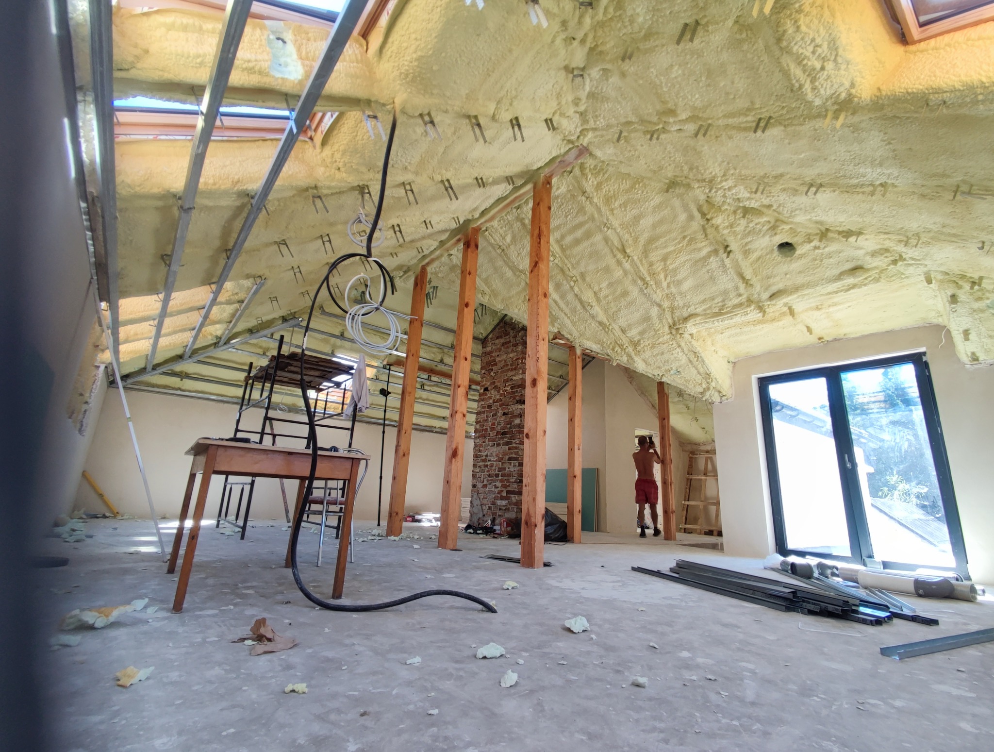 Poddasze w trakcie izolacji pianą PUR. Widoczne drewniane słupy, okna dachowe i fragment muru z cegły. Na podłodze resztki piany i narzędzia.