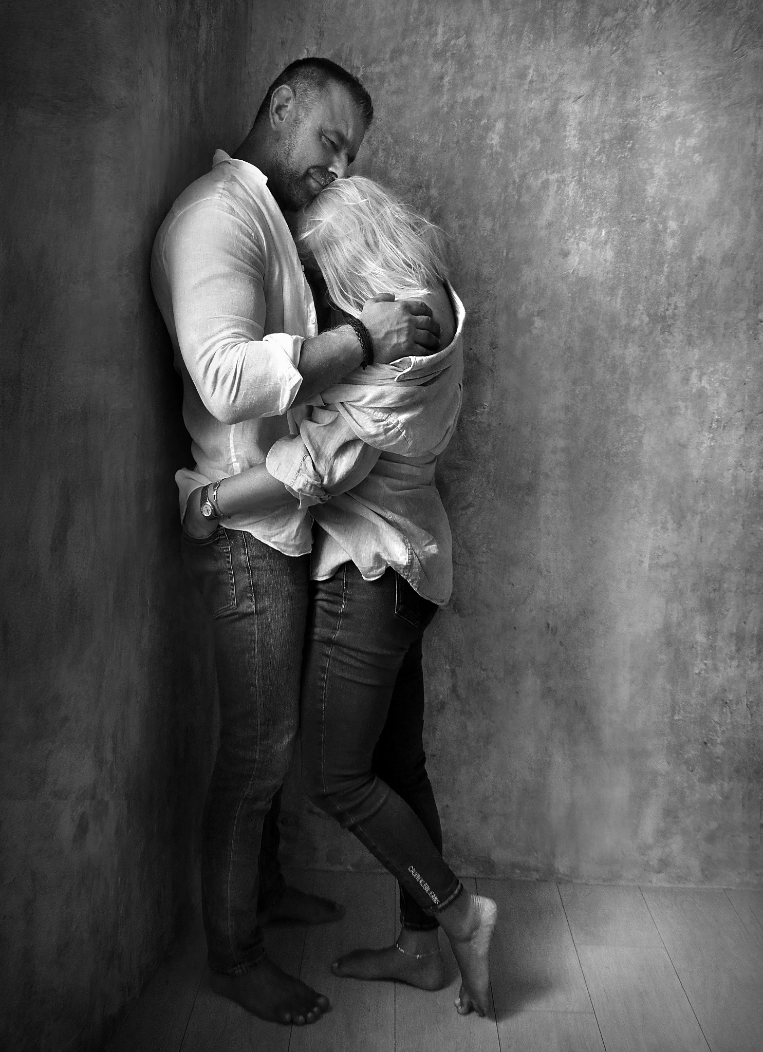 Czarno-białe zdjęcie pary, mężczyzna obejmuje kobietę w białej koszuli i jeansach, stojącej przy szarej ścianie. Widoczne stopy kobiety i fragment podłogi.