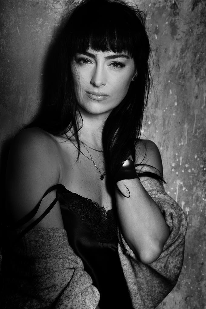 Czarno-biały portret kobiety z grzywką, w bieliźnie z koronką i swetrze, oparta o ścianę z fakturą, z naszyjnikiem. Ujęcie od dołu.