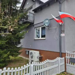 Szary dom z balkonem i białą pompą ciepła York na fundamencie, widoczna zza białego płotu. Na ścianie flaga Polski i antena satelitarna. Numer domu 39.