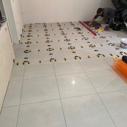 Krzysztof Stożek - Układanie jasnych płytek podłogowych z użyciem systemu poziomowania. Widoczny glazurnik w trakcie pracy, poziomica i narzędzia. W tle wiadro z gruntem.