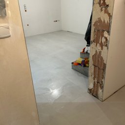 Krzysztof Stożek - Jasnoszara, błyszcząca podłoga z płytek w nowym pomieszczeniu, widok przez futrynę. Fragment ściany z instalacją elektryczną. Narzędzia na schodach w tle.