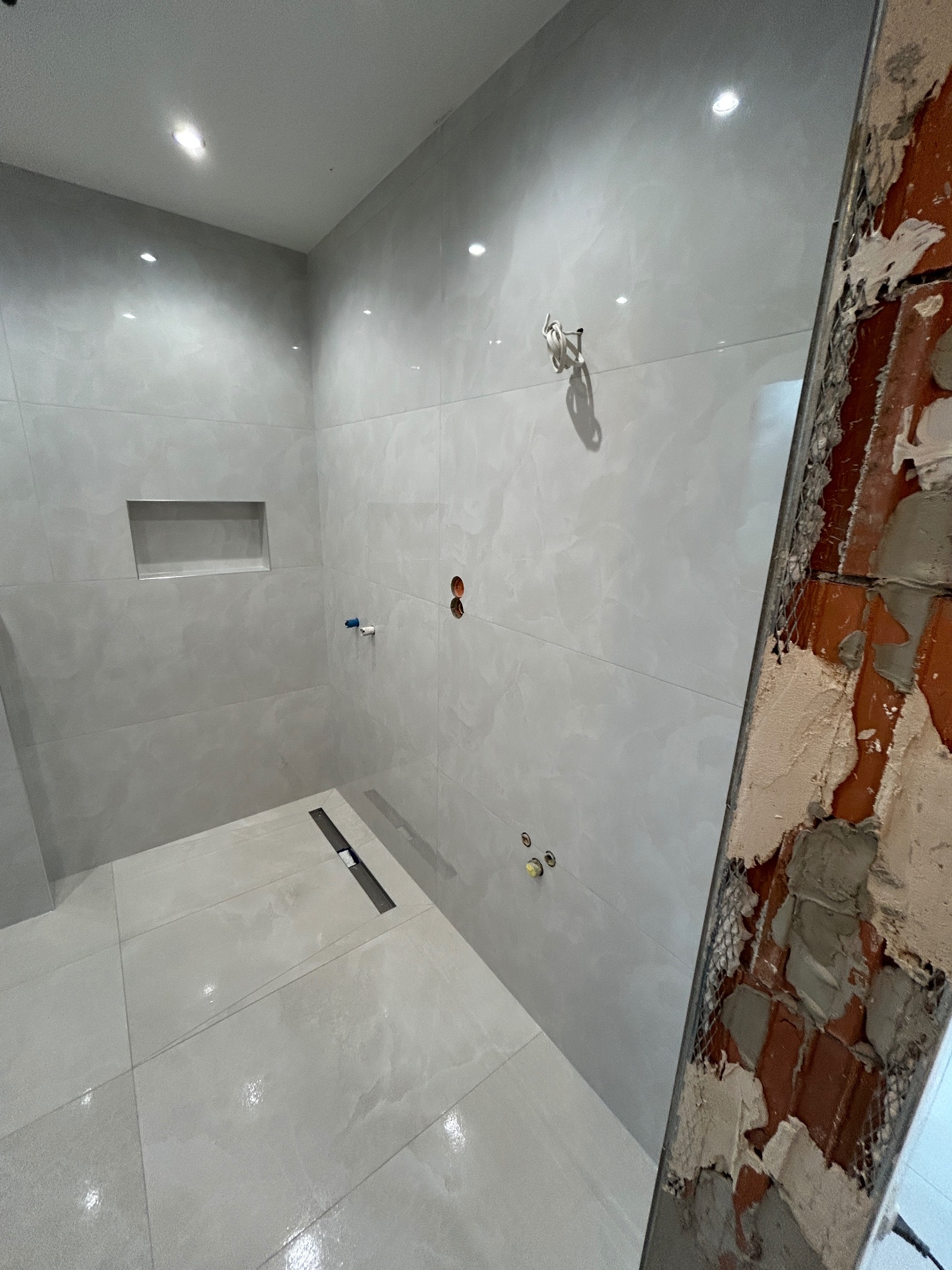 Jasna łazienka w trakcie remontu: świeżo położone płytki podłogowe i ścienne, widoczny odpływ liniowy, fragment ściany w stanie surowym z cegłą i siatką.