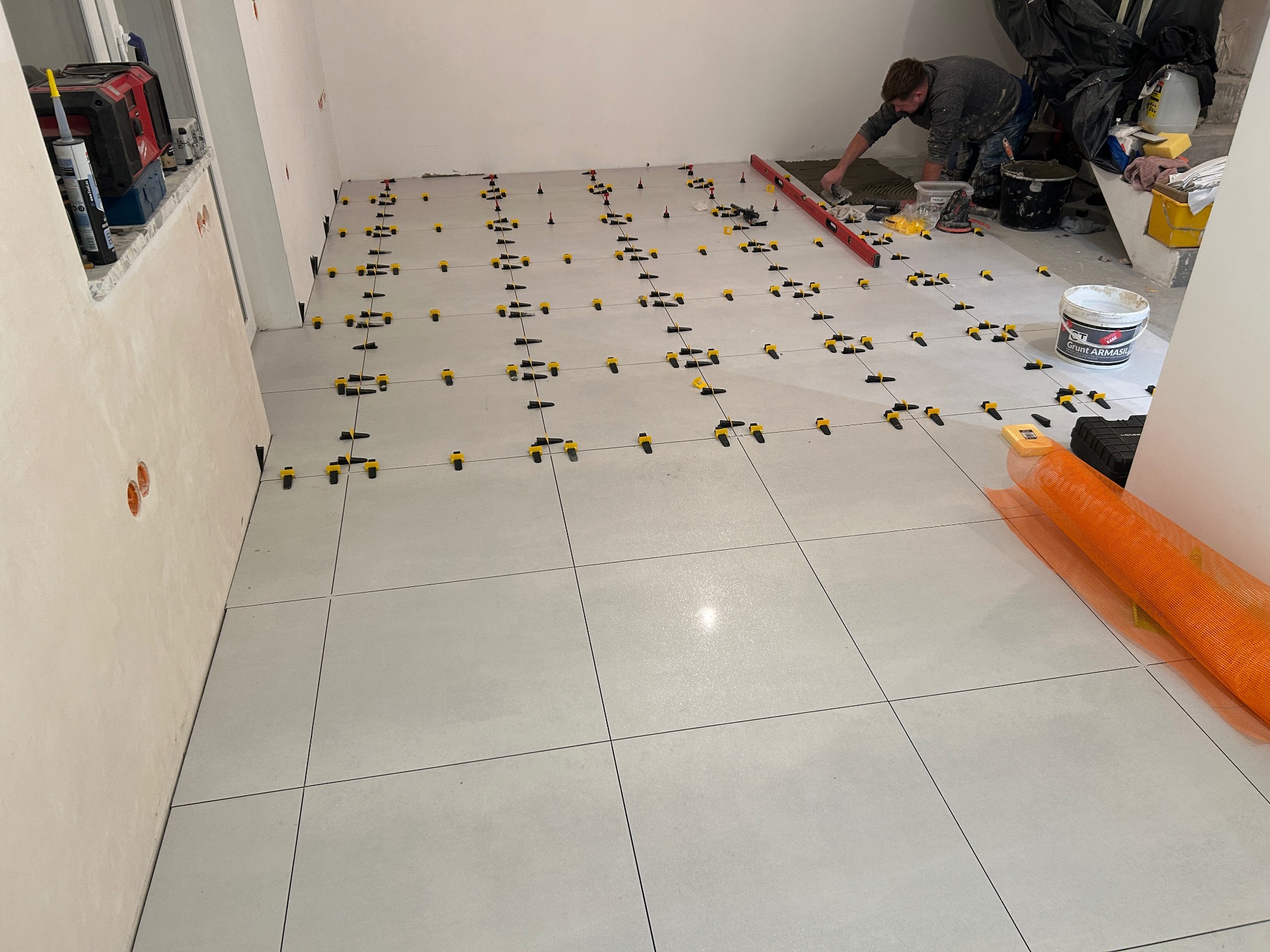 Układanie jasnych płytek podłogowych z użyciem systemu poziomowania. Widoczny glazurnik w trakcie pracy, poziomica i narzędzia. W tle wiadro z gruntem.