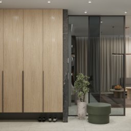 ZMYSŁ WNĘTRZA Architektura Roksana Simińska - Jasna, drewniana szafa wnękowa z długimi, ciemnymi uchwytami, wkomponowana w szarą ścianę z marmuru. Obok szklane drzwi i zielony puf. W tle widok na salon.