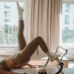 Kobieta w sportowym stroju wykonuje ćwiczenia na reformerze pilates w jasnym studio. Noga uniesiona pionowo, widoczne okna i zasłony w tle.