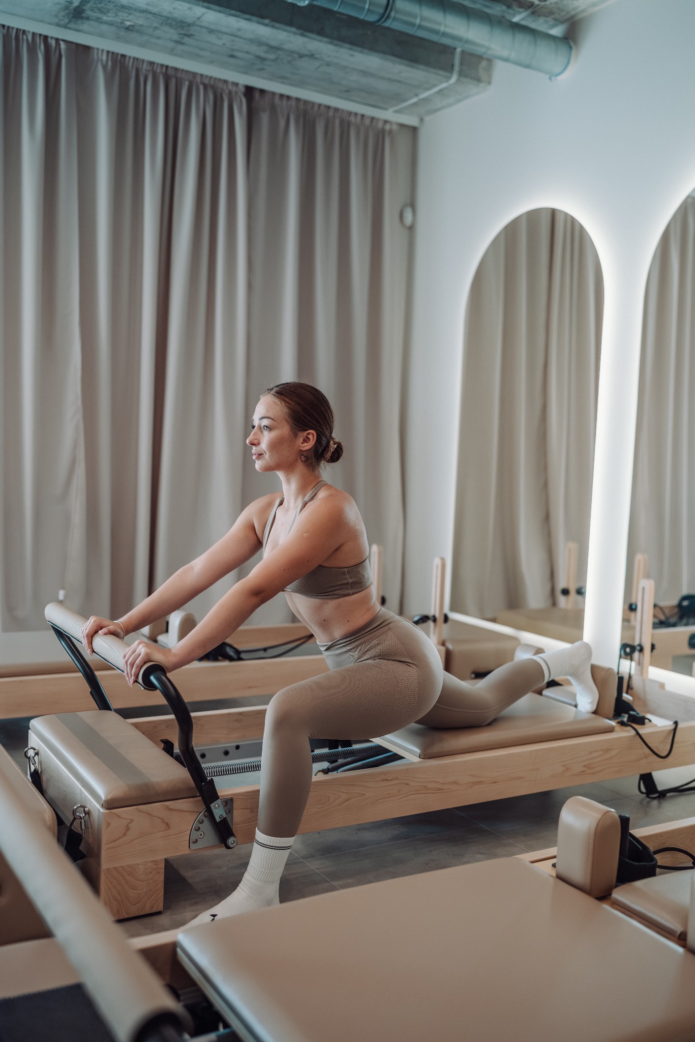 Kobieta w sportowym stroju ćwiczy na reformerze pilates w jasnym studiu z lustrami łukowymi i zasłonami. Skupienie na precyzyjnej pozycji i sprzęcie.