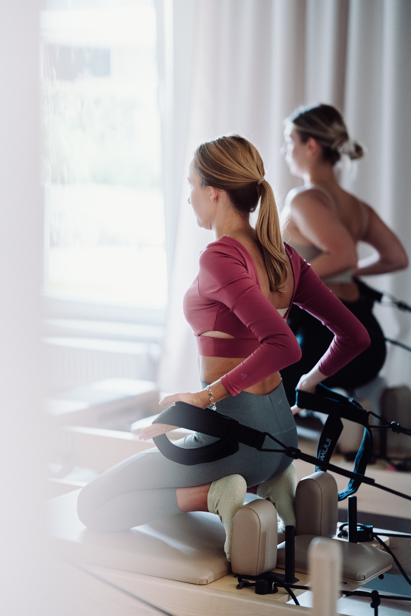 Kobieta w różowej bluzce ćwiczy na reformerze pilates, z tyłu druga osoba w tle. Jasne wnętrze, naturalne światło.