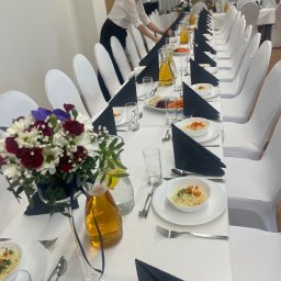 Kamila Dębowska - Uroczysty stół cateringowy z białymi krzesłami, czarnymi serwetkami i kwiatami w wazonie. Osoba w tle przygotowuje stół do przyjęcia.