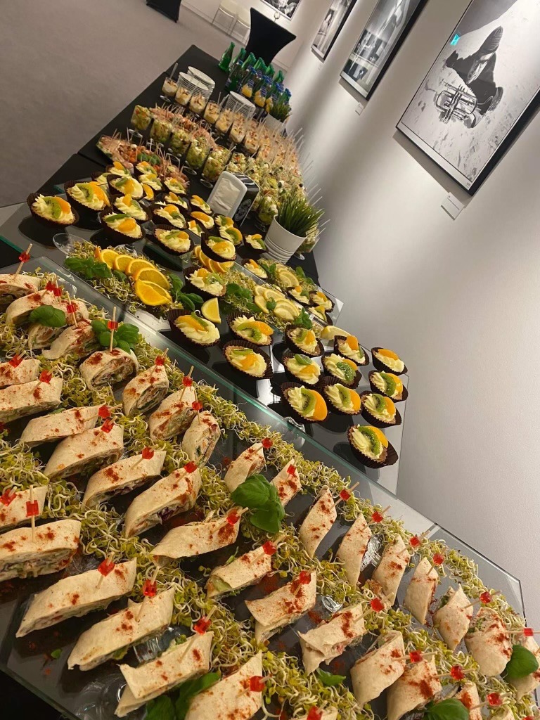 Elegancki stół cateringowy z wrapami, babeczkami z owocami i przekąskami w szklanych naczyniach, udekorowany zieleniną. W tle czarno-białe zdjęcia na ścianie.