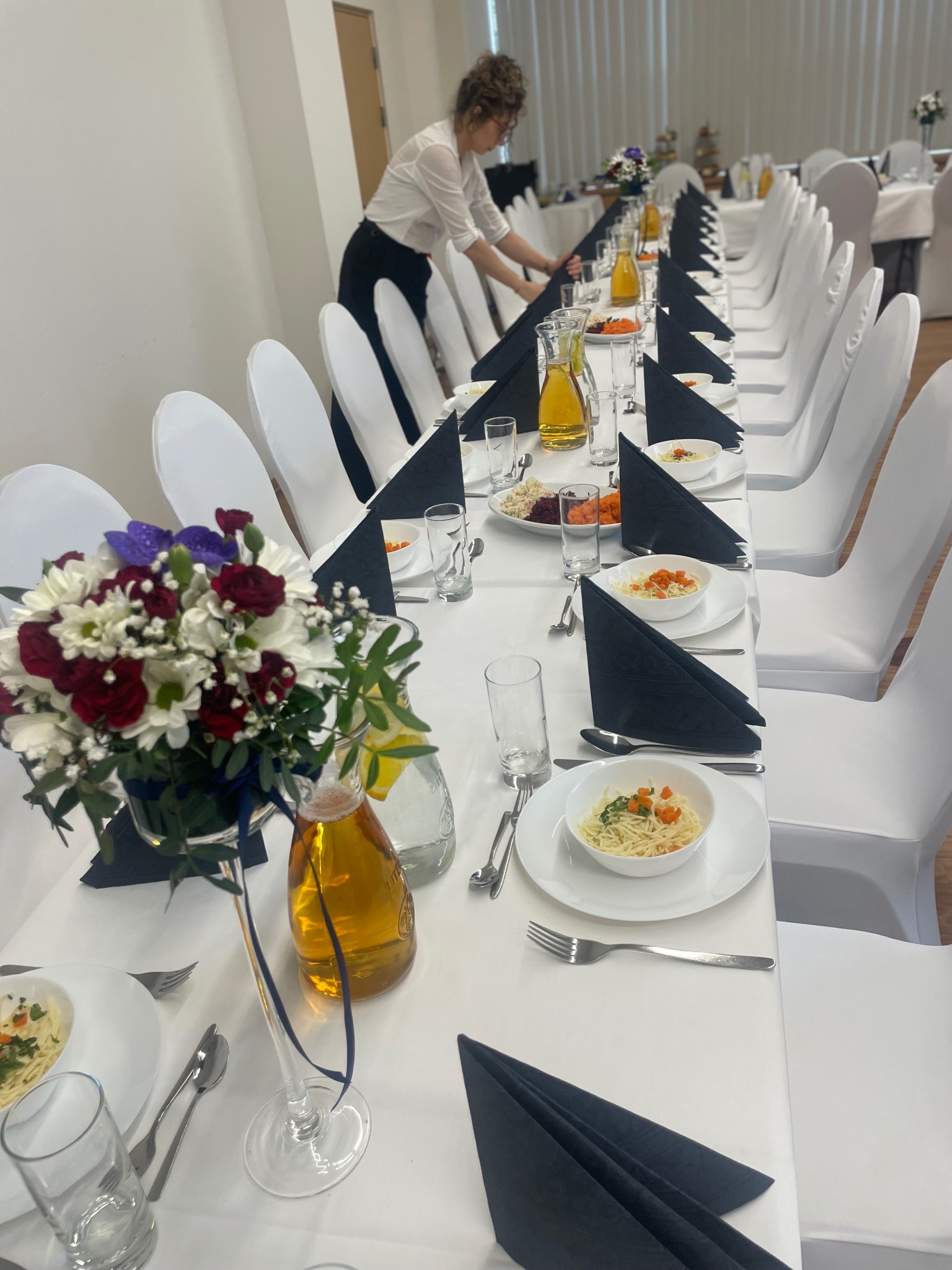 Uroczysty stół cateringowy z białymi krzesłami, czarnymi serwetkami i kwiatami w wazonie. Osoba w tle przygotowuje stół do przyjęcia.
