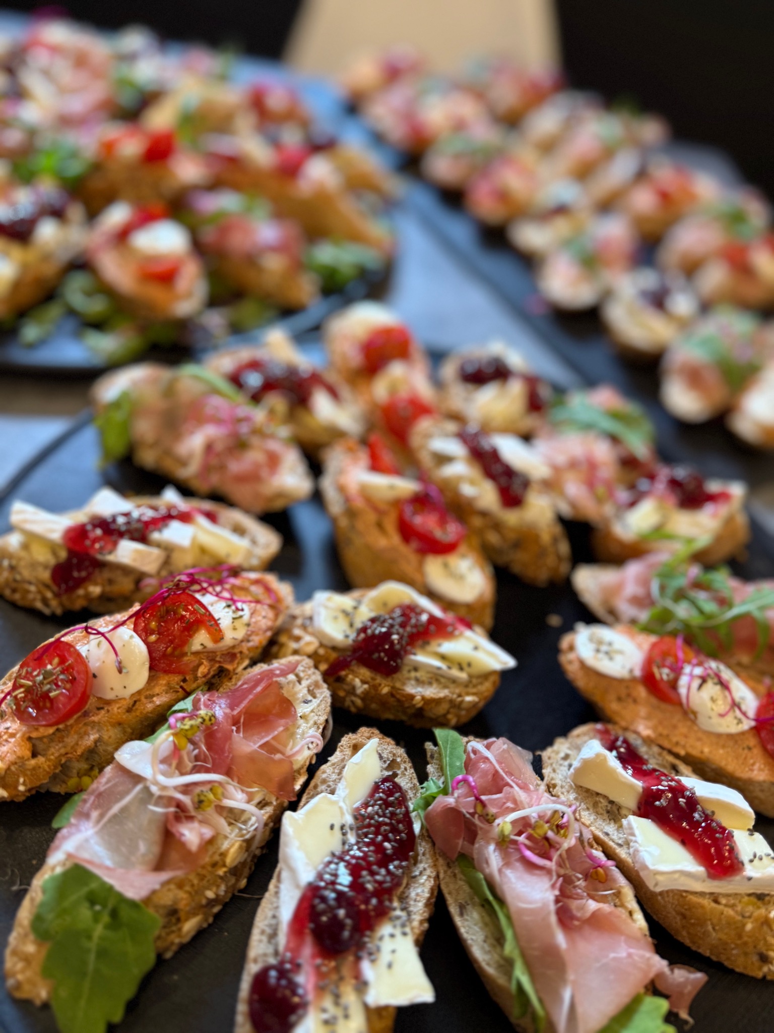 Apetyczne kanapeczki na ciemnym talerzu, dekorowane serem, szynką parmeńską, pomidorkami i dżemem, tworzące elegancki cateringowy zestaw.
