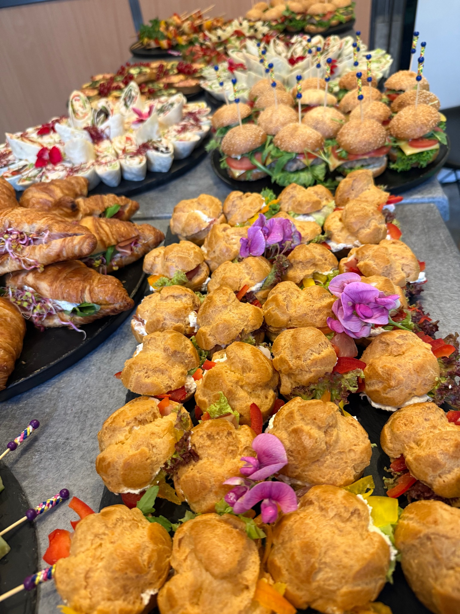 Bogaty wybór przekąsek cateringowych: mini burgery, croissanty z szynką, ptysie z kremem i tortille, elegancko udekorowane kwiatami, na ciemnych tacach.