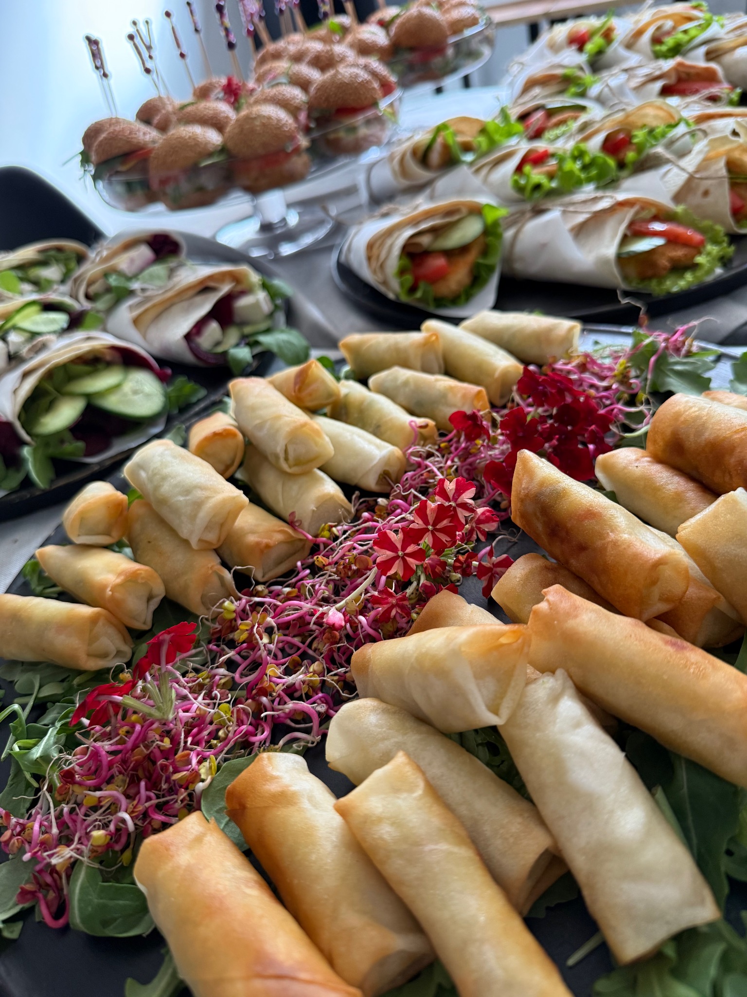 Elegancko podane przekąski cateringowe: mini tortille, sajgonki i mini burgery na czarnych talerzach, udekorowane zielenią i kwiatami. Profesjonalny catering.