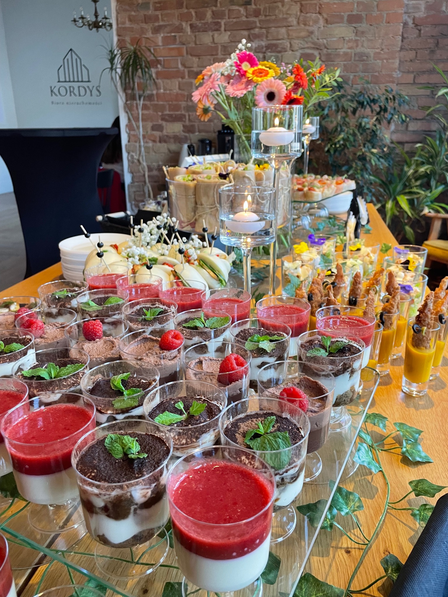 Stół cateringowy z deserami w szklankach, kanapkami i kwiatami, dekorowany zielonymi pnączami. W tle ceglana ściana i logo firmy.