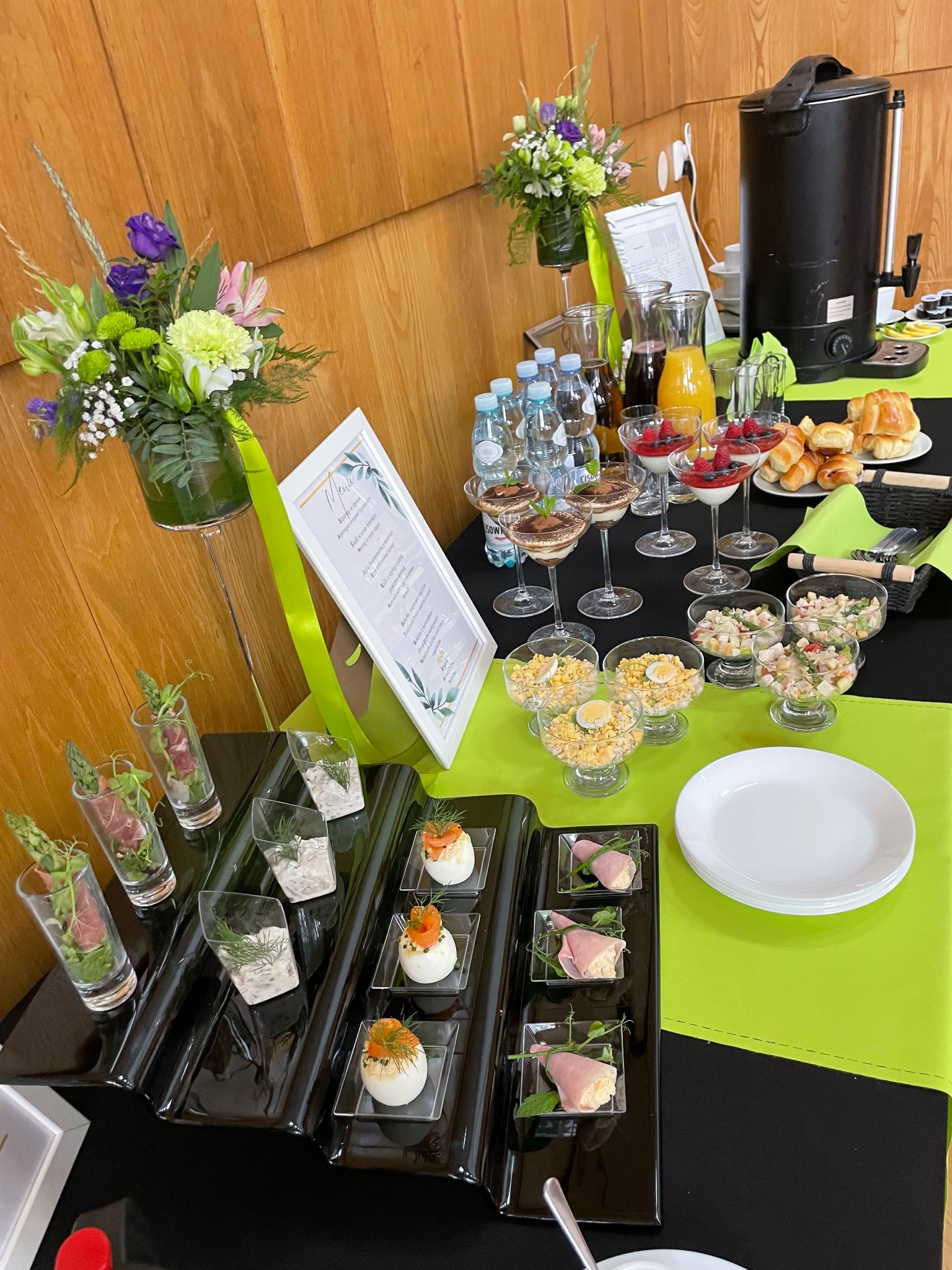 Stół cateringowy z dekoracjami kwiatowymi i różnorodnymi przekąskami: sałatki, desery, jajka faszerowane, rolady z szynki. Bufet w odcieniach czerni, bieli i limonki.