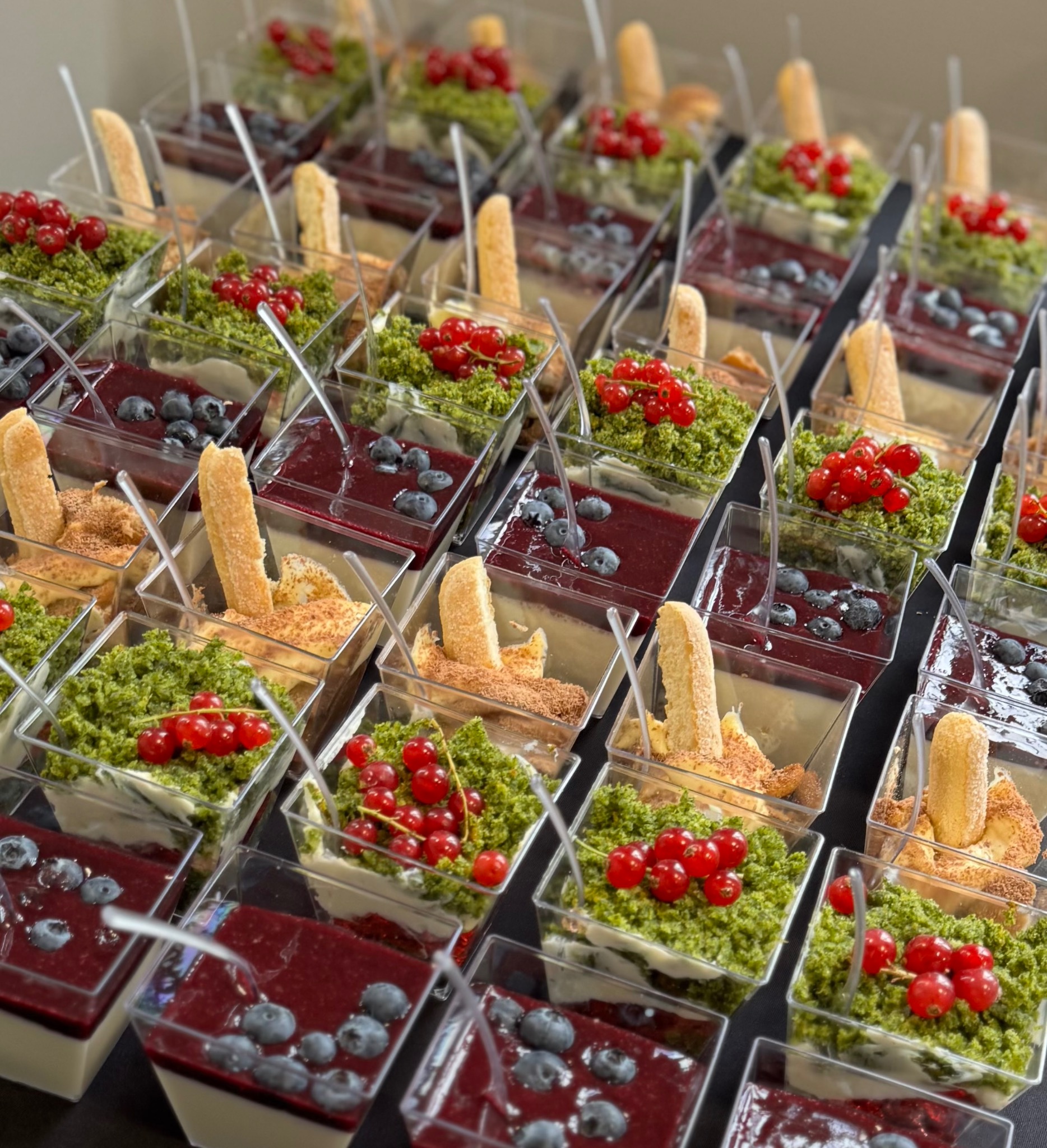 Rzędy deserów w małych, przezroczystych pojemnikach: krem, mus owocowy, posypka i owoce (borówki, porzeczki). Idealne na catering.