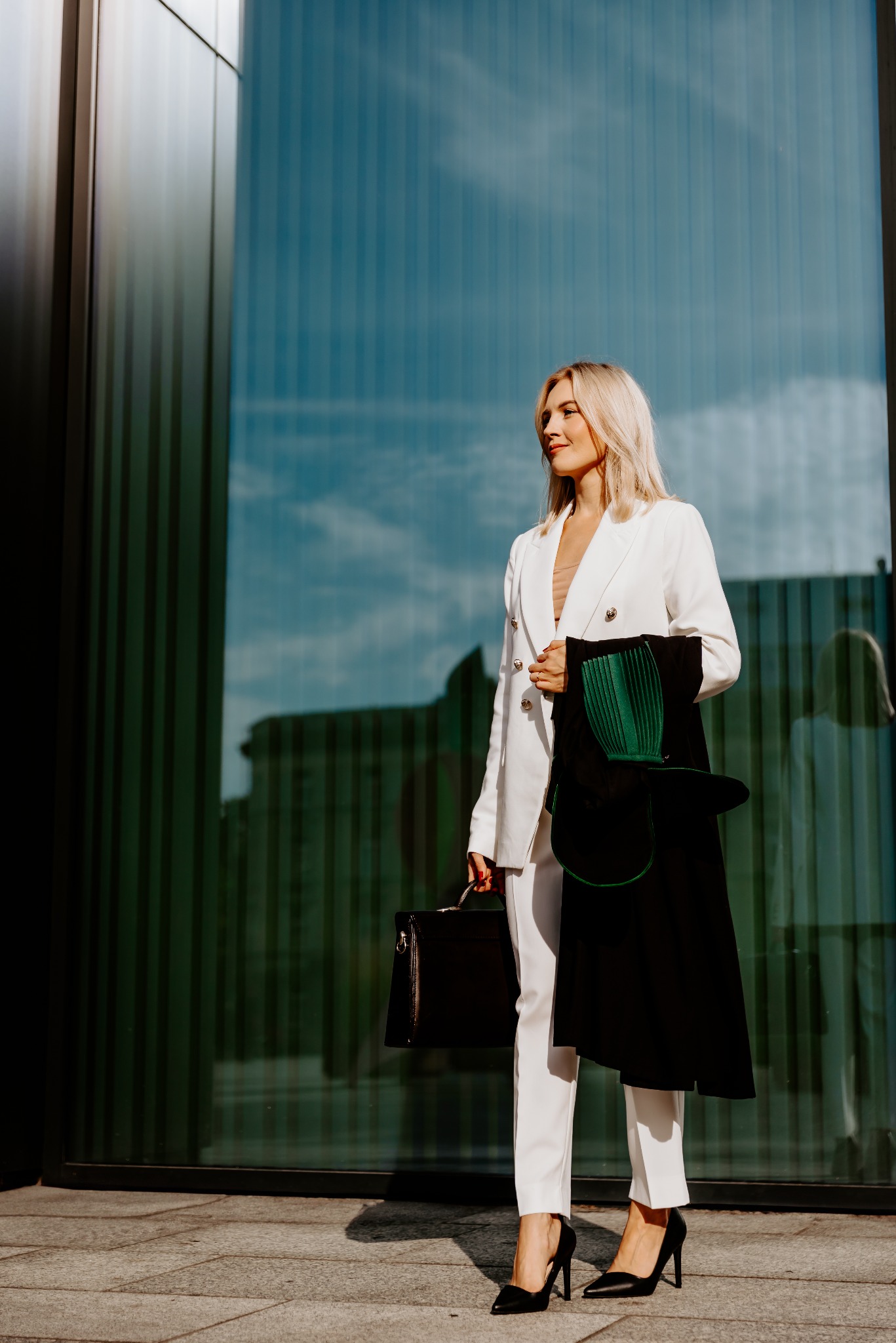 Adwokatka w białym garniturze, trzyma togi i skórzaną aktówkę na tle szklanej fasady budynku, spoglądając w dal. Elegancki i profesjonalny wizerunek.
