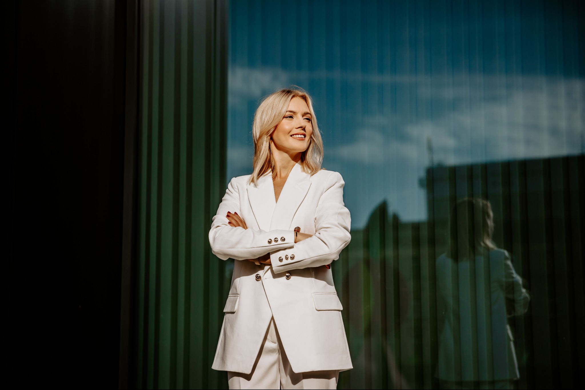 Uśmiechnięta blondynka w białym garniturze na tle nowoczesnej, przeszklonej fasady budynku z odbijającym się niebem.