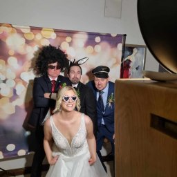 RIVER SPÓŁKA Z OGRANICZONĄ ODPOWIEDZIALNOŚCIĄ - Grupa osób w przebraniach pozuje w fotobudce retro z tłem bokeh, w tym panna młoda w okularach w kształcie serc, peruce afro i uszach królika.