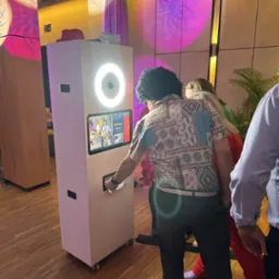 Osoba w peruce odbiera wydruk ze stylowej fotobudki z oświetleniem LED, na tle dekoracji imprezowej. Klasyczna fotobudka na kółkach.