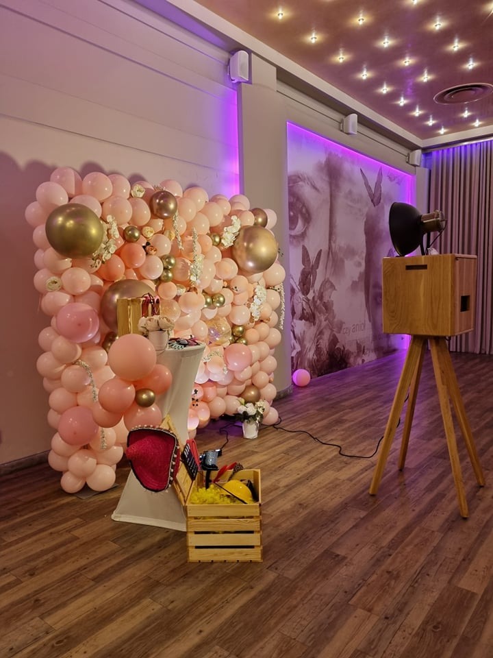 Fotobudka w stylu retro z dekoracją z różowych i złotych balonów, drewnianą obudową i rekwizytami w skrzynce. W tle grafika na ścianie i oświetlenie punktowe.