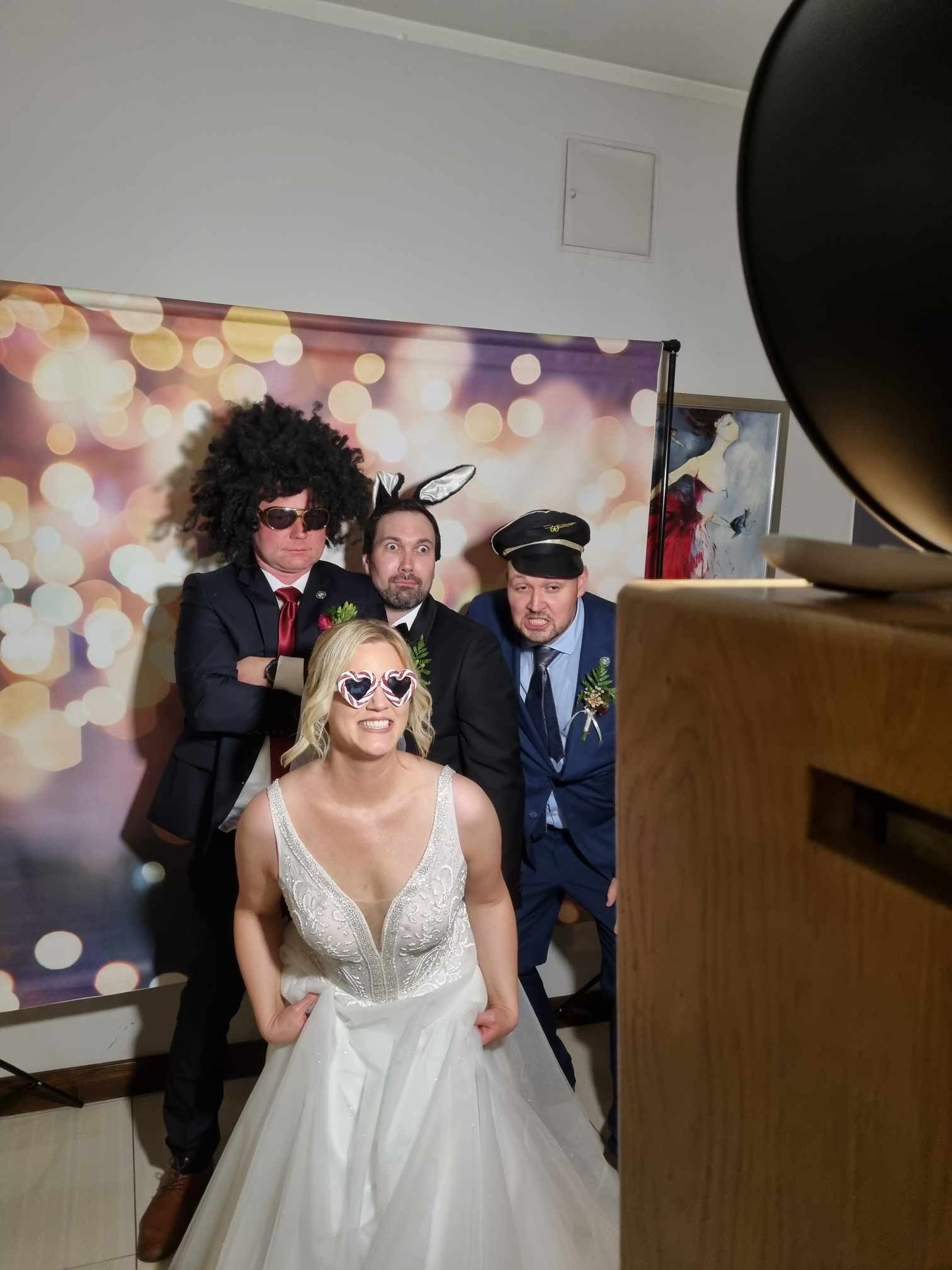 Grupa osób w przebraniach pozuje w fotobudce retro z tłem bokeh, w tym panna młoda w okularach w kształcie serc, peruce afro i uszach królika.