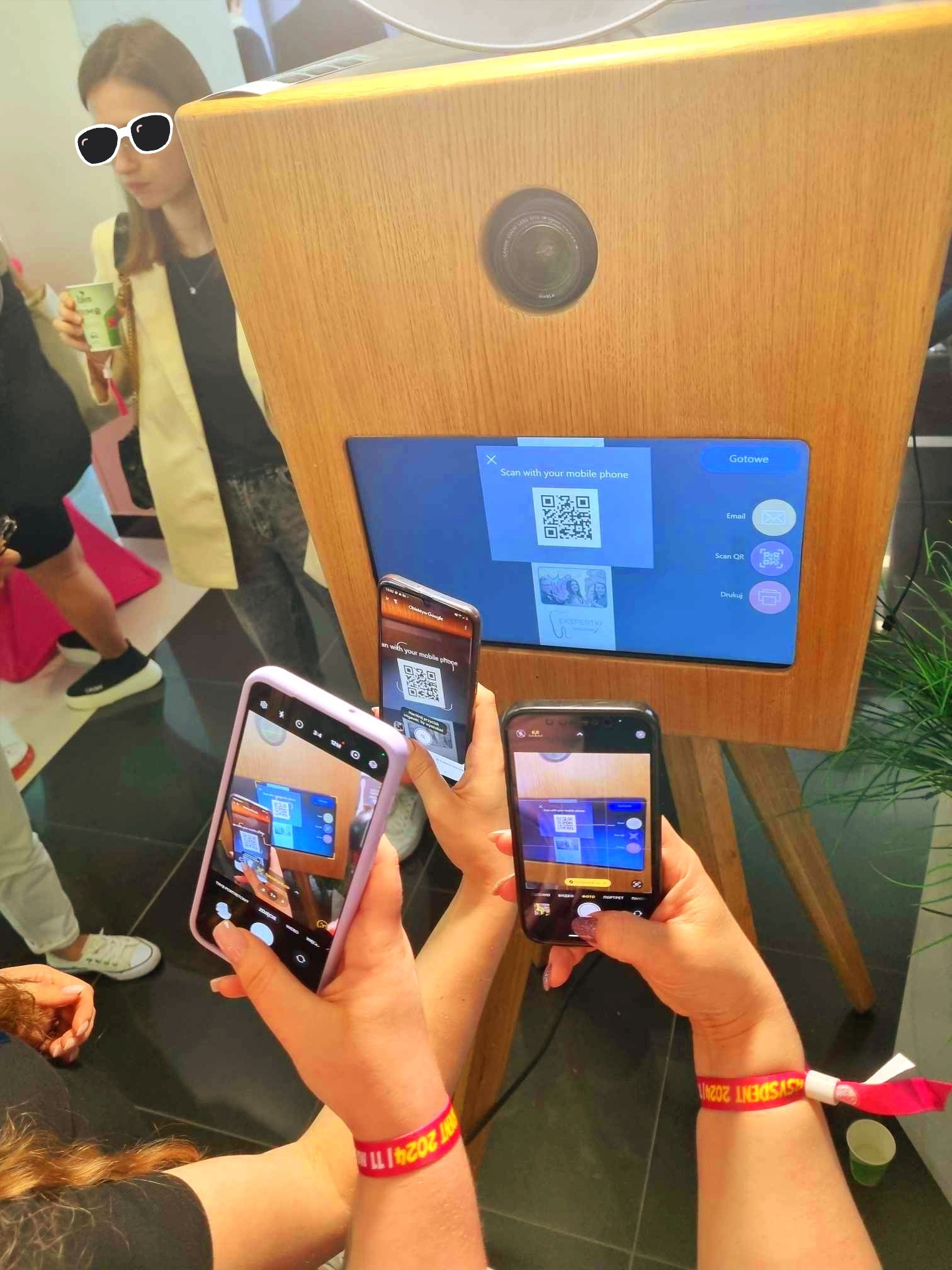 Osoby skanują kod QR na ekranie fotobudki retro za pomocą telefonów komórkowych. Widoczne opcje udostępniania: email, skan QR, druk.