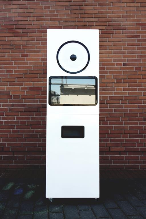 Biała, klasyczna fotobudka na kółkach, z okrągłym obiektywem i ekranem podglądu, stoi na kostce brukowej przed ceglaną ścianą. Widoczne odbicie budynku w szybie.