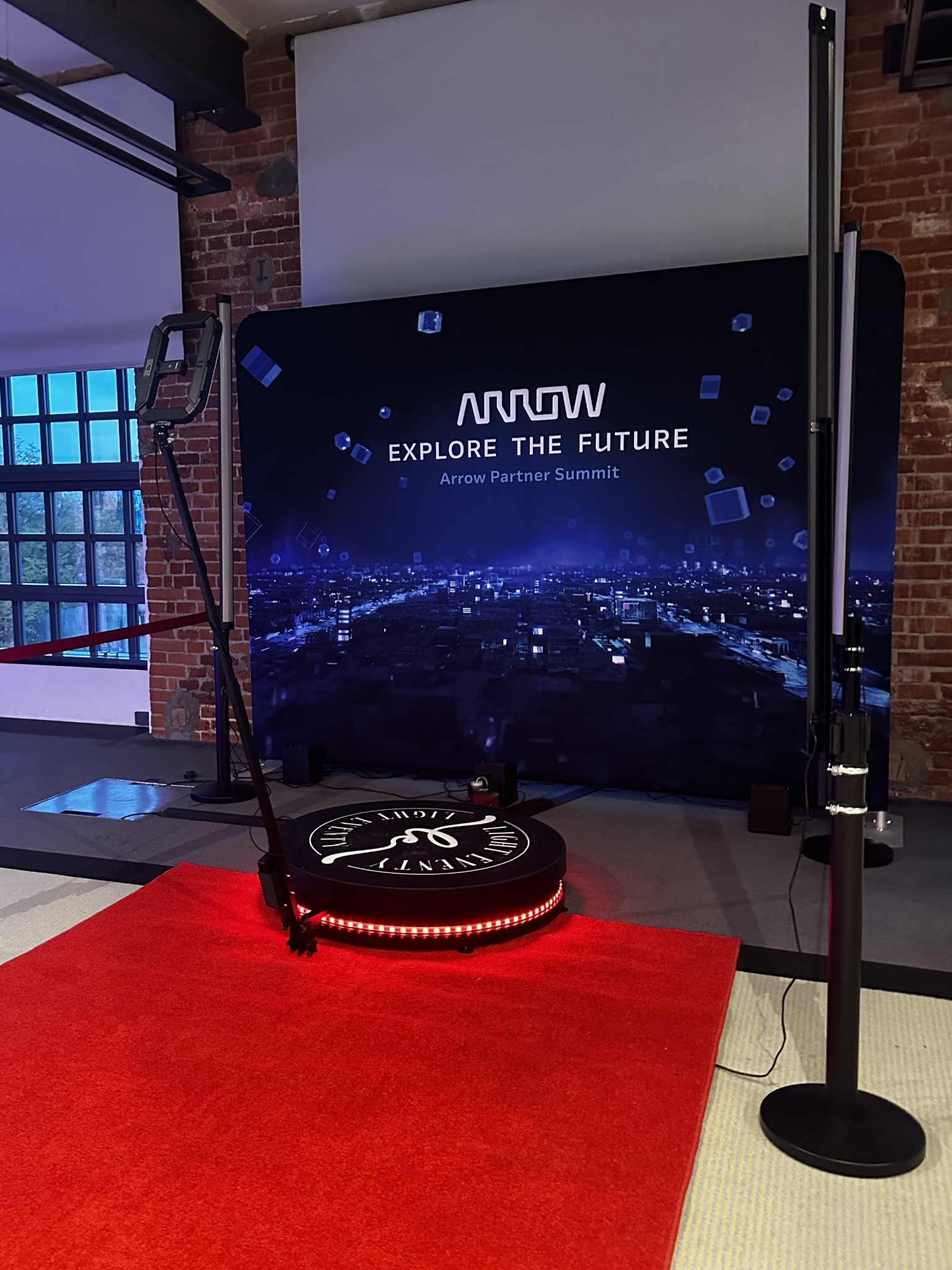 Fotobudka 360 z platformą na czerwonym dywanie i tłem z grafiką miejską, ustawiona w industrialnym wnętrzu z ceglanymi ścianami i oknem.