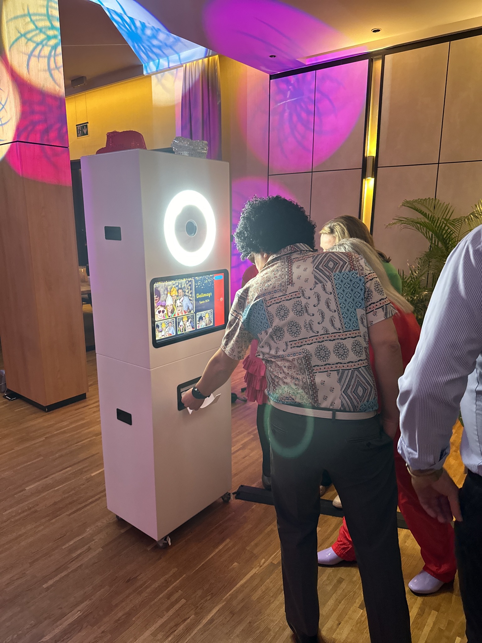 Osoba w peruce odbiera wydruk ze stylowej fotobudki z oświetleniem LED, na tle dekoracji imprezowej. Klasyczna fotobudka na kółkach.