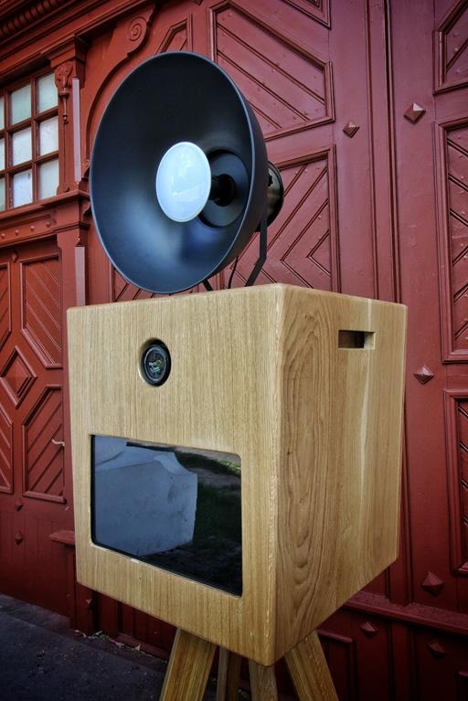 Retro fotobudka z drewnianą obudową i lampą w stylu vintage, na tle czerwonych, zdobionych drzwi. Widoczny ekran i elementy konstrukcyjne.