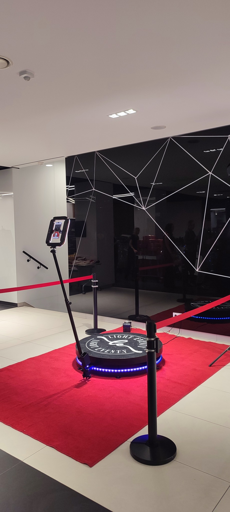 Rotacyjna platforma z ekranem i oświetleniem LED na czerwonym dywanie, otoczona barierkami. Czarno-białe tło z geometrycznym wzorem. Nowoczesny design.