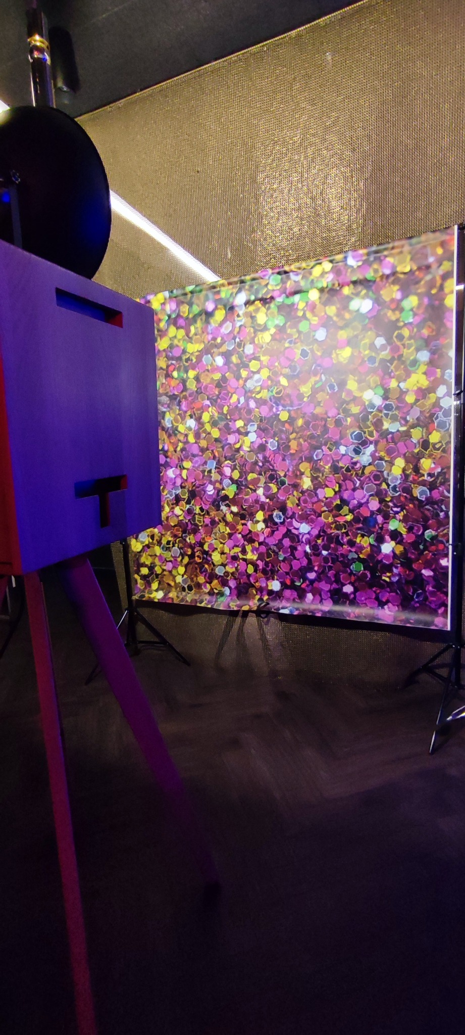 Fioletowa fotobudka z tłem w kolorowe cekiny, ustawiona na drewnianej podłodze, gotowa do użycia podczas imprezy. Oświetlenie podkreśla błyszczące tło.