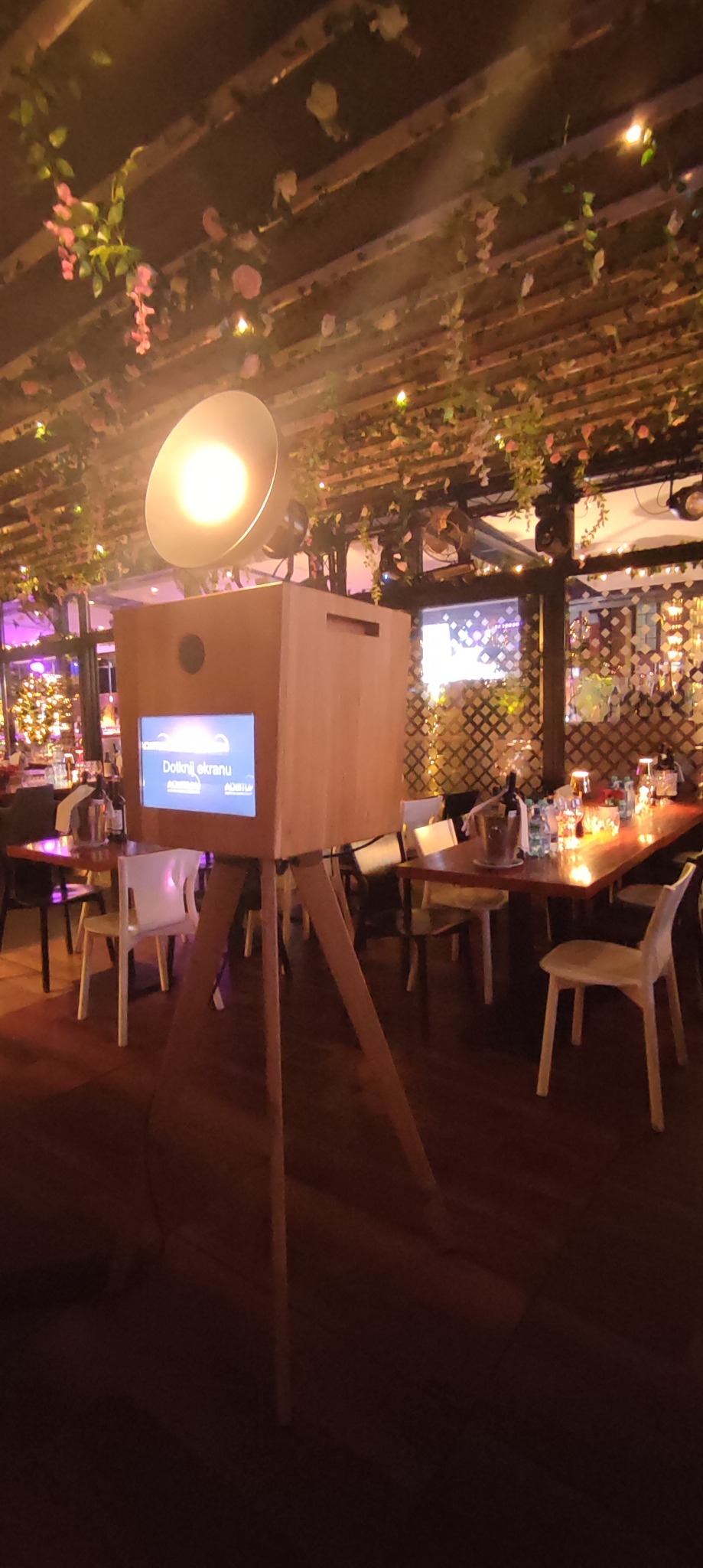 Drewniana fotobudka z lampą na trójnogu, ustawiona w udekorowanym kwiatami wnętrzu restauracji, gotowa do użycia podczas imprezy.