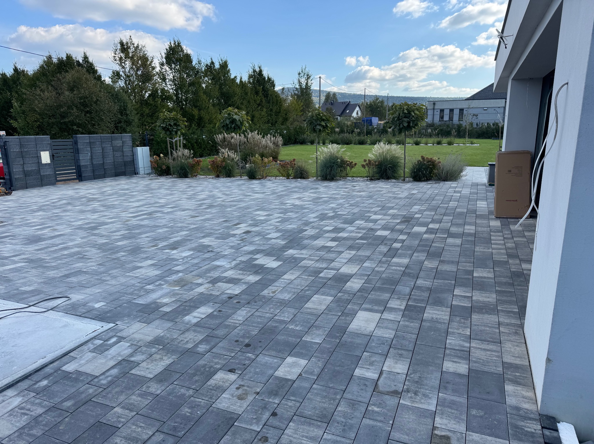 Ułożona, szara kostka brukowa na podjeździe domu jednorodzinnego. Widoczny fragment ogrodu z ozdobnymi trawami i krzewami. Nowoczesne ogrodzenie panelowe w tle.