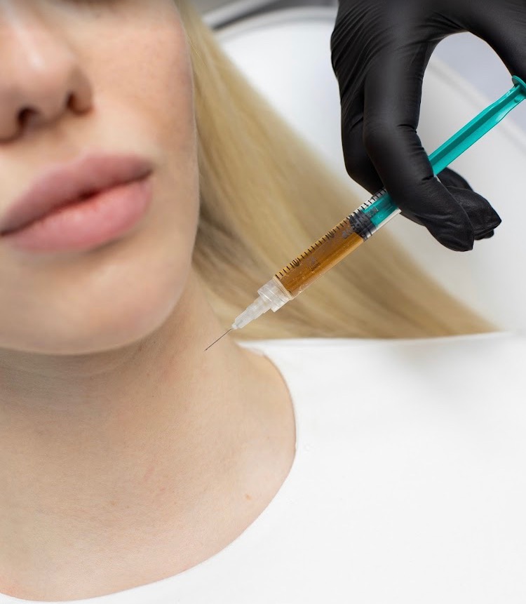 Iniekcja w szyję pacjentki z blond włosami, wykonywana przez osobę w czarnych rękawiczkach, z użyciem strzykawki z zielonym tłokiem i brązowawą substancją.