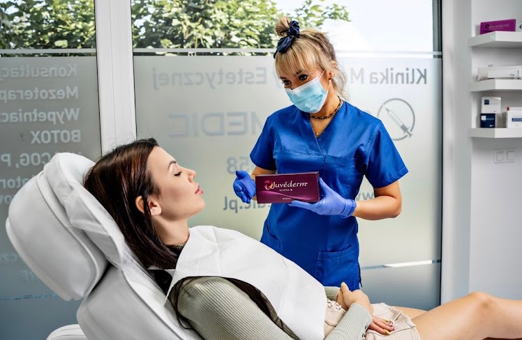 Klientka leży na fotelu, a lekarz w niebieskim uniformie trzyma w dłoniach preparat Juvederm. Widoczne logo kliniki na szybie, półki z produktami w tle.